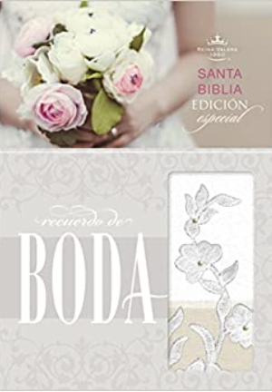 Una mujer sostiene un ramo de flores blancas y rosas sobre una «Biblia de boda» decorativa con bordados florales y la inscripción «recuerdo de boda».
