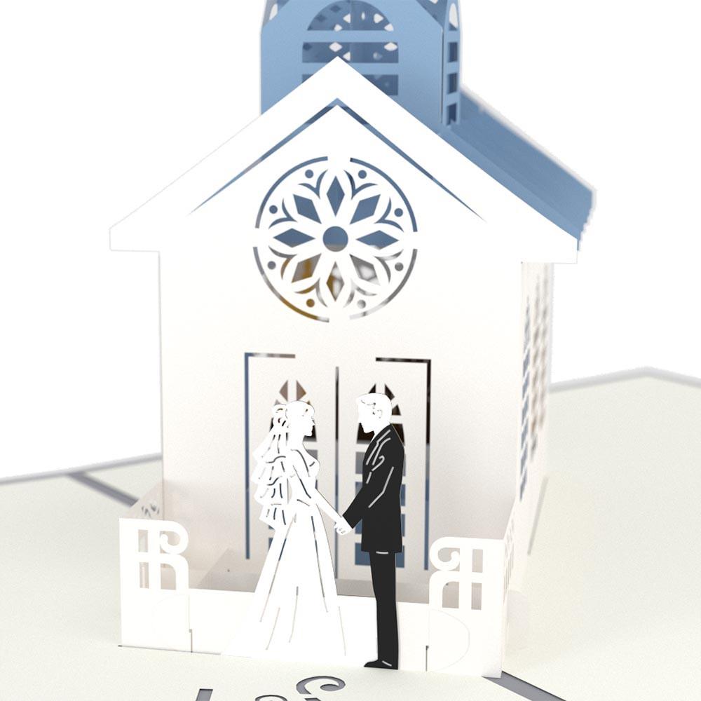 Esta tarjeta de alta calidad es una tarjeta pop-up de 5 x 7 en la que aparecen unos novios de pie frente a una capilla blanca con techo azul y una ventana circular decorativa.