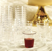 Un grupo de vasos de plástico transparentes para la comunión apilados, con un vaso en primer plano que contiene un líquido rojo, sobre un mantel blanco estampado: perfectos para tu próxima compra o si necesitas una caja de 1000 unidades.